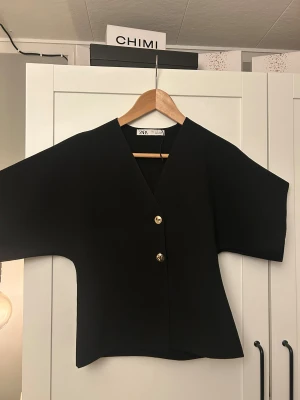 Svart kofta med guldfärgade knappar från Zara - Stilren svart kofta från Zara i storlek S med v-ringad hals och två guldfärgade knappar framtill. Koftan har breda, korta ärmar och är tillverkad i ett mjukt stickat material som ger en skön känsla. 