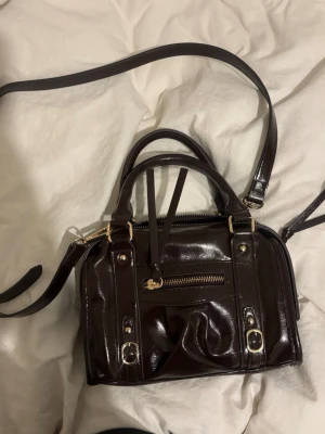 Mörkbrun handväska med gulddetaljer - Snygg mörkbrun handväska i glansigt skinn med både axelrem och handtag. Väskan har guldfärgade detaljer, dragkedja framtill och dekorativa spännen. Perfekt storlek för att få plats med allt viktigt och passar till många olika stilar.