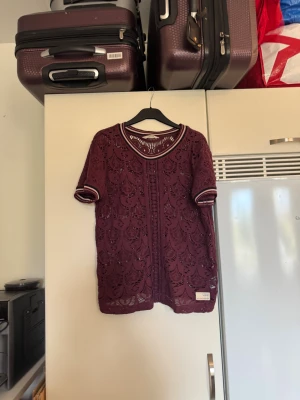 Vinröd spetsblus med sportiga detaljer - Säljer en vinröd blus med korta ärmar och sportiga ribbade muddar i vinrött, rosa och svart. Blusen är helspets med ett snyggt mönster och har rund halsringning med samma ribbade kant som vid ärmsluten. Perfekt för dig som gillar att sticka ut med detaljer. Från märket Odd Molly. Storlek 2 som enligt deras storleksguide motsvarar en M. Mycket fint skick, jag hittar inga synliga anmärkningar eller defekter.  