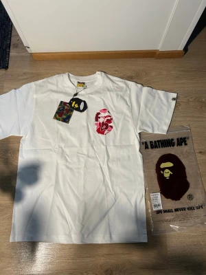 Vit BAPE x Stüssy t-shirt M - Vit t-shirt från BAPE x Stüssy med ikoniskt ape-head tryck i röd och rosa camo på bröstet och ryggen. Stora röda bokstäver och Stüssy-logga på ryggen. Klassisk rund hals och korta ärmar. Märkt med BAPE-tag på ärmen. Skön bomullskvalitet.
