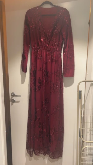 Vinröd långklänning med paljetter - Vinröd super snygg långklänning med v-ringning och långa ärmar. Klänningen har glittriga paljetter i blommigt mönster över hela tyget. Älskar verkligen denna klänning men den är tyvärr för stor för mig, finns en liten ”reva” på klänningen längst ner men syns inte när den är på (se bild)