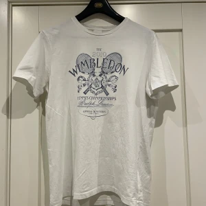 Vit Wimbledon t-shirt från Polo Ralph Lauren - Vit t-shirt från Polo Ralph Lauren med Wimbledon 2010-tryck i blått framtill. Klassisk rund hals och korta ärmar. Tillverkad i 100% bomull för skön och mjuk känsla. Perfekt för dig som gillar tennis och sportig stil.