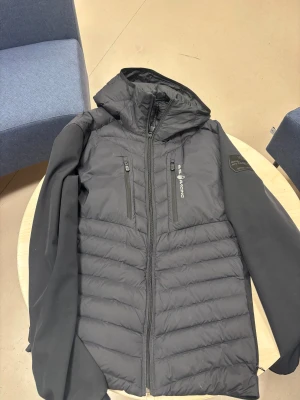 Svart pufferjacka från Sail Racing - Svart pufferjacka från Sail Racing med huva och två vertikala bröstfickor med dragkedja. Jackan har quiltad framsida och baksida, samt släta svarta ärmar. Materialet är polyester och syntet, perfekt för kyliga dagar. Snygg logga på bröstet och ärmen. Äkta