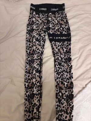 Leopardmönstrade leggings från Stronger - Snygga leggings från Stronger med leopardmönster i svart, beige och bruna toner. De har en bred svart midja med loggan STRONGER tryckt runtom. Perfekta för träning och har en tight passform som sitter skönt mot kroppen.