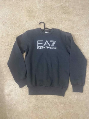 Mörkgrå sweatshirt EA7 Emporio Armani - Snygg mörkgrå sweatshirt från EA7 Emporio Armani med stort vitt tryck på bröstet. Tröjan har rund hals, långa ärmar och ribbade muddar vid ärmslut och nederkant. Perfekt för dig som gillar stilrena märkesplagg med sportig vibe.