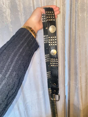 Svart skinnväst med nitar och mynt - Cool svart skärp i skinn med massor av silvriga nitar och stora myntdetaljer med kronmotiv. Skärpet har ett brett band och metallspänne. Perfekt för dig som gillar edgy och unik stil med detaljer som sticker ut.