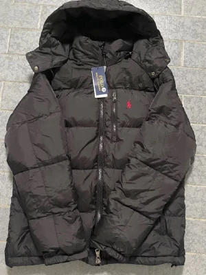 Svart pufferjacka från Polo Ralph Lauren - Svart pufferjacka från Polo Ralph Lauren med huva och dragkedja framtill. Jackan har två sidofickor med dragkedja och en röd Polo-logga på bröstet. Materialet är syntet och jackan har en quiltad design som ger extra värme. Perfekt för kalla dagar. Har i storlek S oxå👍🏼