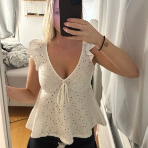 Vit blus med volangärmar och knyt - Supersöt vit blus i bomull med broderade hål och volangärmar. Djup v-ringning och knyt framtill ger en romantisk vibe. Perfekt för varma dagar och somriga outfits.