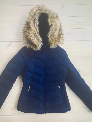 Tessie vinterjacka stl XS/32 - Snygg mörkblå pufferjacka från Tessie i Göteborg med fluffig huva kantad av beige fuskpäls. Jackan har dragkedja framtill och två fickor med dragkedja. Perfekt för kalla dagar och har en schysst figurnära passform.