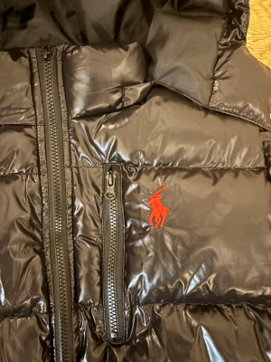 Svart pufferjacka från Polo Ralph Lauren - Säljer en svart pufferjacka från Polo Ralph Lauren med glansig finish och röd broderad logga på bröstet. Jackan har huva, flera dragkedjor och är långärmad. Perfekt för kalla dagar och har en riktigt schysst streetstil.