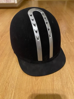 Svart ridhjälm med silverdetaljer - Säljer en svart ridhjälm från EQUESTRIAN HELMETS i storlek 57. Hjälmen har två snygga silverfärgade detaljer på ovansidan och en klassisk skärm framtill. Insidan är vadderad med mesh för extra komfort och ventilation.