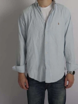 Ljusblå skjorta från Ralph Lauren - Säljer en klassisk ljusblå skjorta från Ralph Lauren med button-down krage och broderad logga på bröstet. Skjortan har långa ärmar och knappar framtill. Perfekt för dig som gillar stilrena och tidlösa plagg.