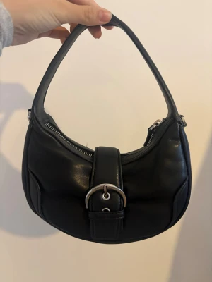 Svart axelväska med silverdetalj - Snygg svart axelväska i skinn med rundad form och ett brett axelband. Väskan har en dekorativ silverfärgad spänne framtill och dragkedja upptill. Perfekt storlek för det viktigaste och passar till många olika stilar.