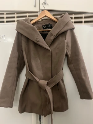 Beige kappa med huva från Newyorker (Amisu) - Snygg beige kappa från Amisu i storlek 34 med bred huva och knytskärp i midjan. Jackan har långa ärmar, knappar framtill och markerad midja för en feminin siluett. Perfekt för dig som gillar stilrena och klassiska plagg.