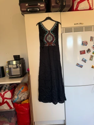 Svart långklänning med broderi - Svart långklänning med färgstarkt broderat mönster i lila, turkos, orange och vitt vid bröstet. Klänningen har v-ringad hals och är ärmlös, med ett mjukt och lätt tyg som ger ett flowy intryck. Perfekt för dig som gillar boho-vibe och detaljer. Från märket Odd Molly. Storlek 0 som enligt deras storleksguide motsvarar en XS. Men passar en  S. Helt ny med tags/ prislapp fortfarande kvar. 