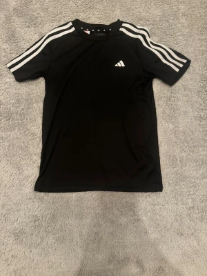 Svart Adidas t-shirt med vita ränder - Svart t-shirt från Adidas med klassiska vita ränder längs axlarna och ärmarna. Adidas-logga på bröstet och rund hals. Tillverkad i lätt polyester med AEROREADY-teknologi som håller dig fräsch under träning. Perfekt för sport eller chill.