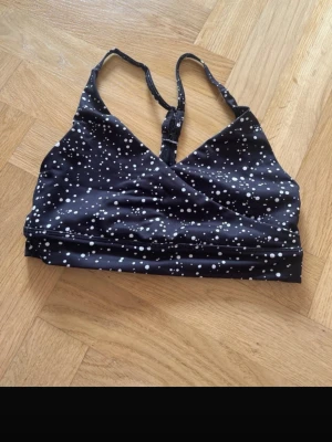 Svart sport-bh Aim'n med prickigt mönster - Svart sport-bh från Aim'n med vitt prickigt mönster. Bh:n har smala axelband som går ihop i ryggen och är tillverkad i stretchigt material med 73% polyester och 27% spandex. Perfekt för träning och har en snygg omlott-design framtill.