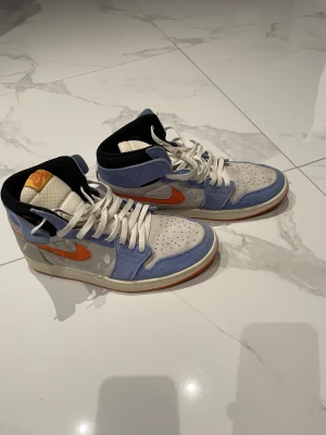 Nike Jordan 1 High blå och orange - Nike Jordan 1 High sneakers med vit textil och blå mocka-paneler, orange swoosh och detaljer. Skorna har snörning, perforerad tå och svart foder. Tungan har en gul etikett med röd 'Zoom Air'-logga. Snygg kontrast mellan färgerna och klassisk siluett.