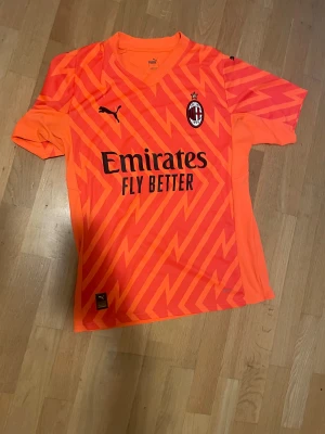 Inga skor på bilderna - Bilderna visar en orange AC Milan fotbollströja från Puma med zigzag-mönster och sponsortryck, men inga skor finns med.