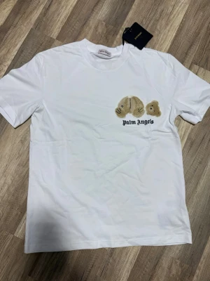 Vit Palm Angels t-shirt - Vit t-shirt från Palm Angels med ikoniskt nallebroderi på bröstet och svart logotyp under. Klassisk passform och rund halsringning. 