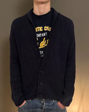 Mörkblå ribbstickad kofta - Säljer en mörkblå ribbstickad kofta med knappar framtill och bred krage. Koftan har lång ärm och en klassisk, lite loose passform. Perfekt att slänga på sig över en t-shirt för en chill look.