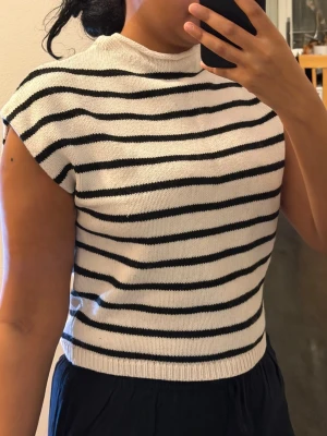 Randig stickad ärmlös topp - Snygg ärmlös topp med svartvita horisontella ränder och ribbad kant nertill. Toppen har en högre halsringning och är stickad i mjukt material, perfekt för en chill och stilren look.