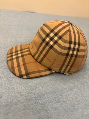 Rutig Burberry keps i beige och brun - Snygg Burberry keps med klassiskt rutigt mönster i beige, brunt och svart med röda detaljer. Justerbar baktill med svart plastspänne och tydlig Burberry London England patch. Materialet är bomull och polyester, perfekt för dig som gillar ikoniska accessoarer.