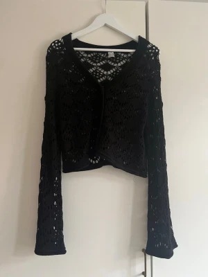 Svart virkad kofta från H&M Divided - Säljer en svart, virkad kofta från H&M Divided i storlek S. Koftan har långärmade ärmar och ett snyggt genombrutet mönster som ger en cool och trendig vibe. Perfekt att slänga över en topp för en chill look. Croppad passform och mjukt stickat material.