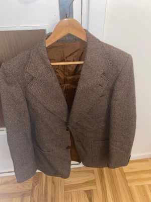 Brun kavaj från Vinson Strands - Snygg brun kavaj från Vinson Strands med klassisk krage och två framfickor. Fodret är i glansig brun polyester och jackan har en diskret rutig struktur. Perfekt för dig som vill ha en stilren och tidlös look.