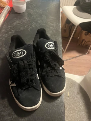 Adidas Campus svarta sneakers - Svarta Adidas Campus sneakers med vita ränder och logga på plösen. Skorna har snörning och en platt, gummisula i orange. Ovandelen är i mocka och insidan är mjuk. Klassisk och stilren modell med rund tå och låg profil. Inte super använda så dem är i bra skick. 