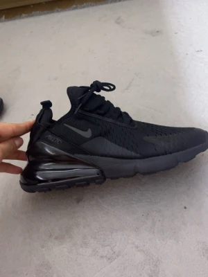 Nike Air Max 270 Svarta sneakers - Säljer ett par svarta Nike Air Max 270 sneakers med snygg, sportig design och synlig luftkudde i hälen. Skorna har mesh-ovandel, snörning och platt sula. Perfekta för dig som gillar streetstyle och vill ha bekväma skor med grym dämpning.