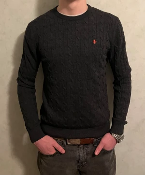 Mörkgrå stickad tröja från Morris - Säljer en mörkgrå kabelstickad tröja från Morris i 100% merinoull. Tröjan har rund hals, lång ärm och en liten röd broderad logga på bröstet. Perfekt för dig som gillar klassisk stil med snygga detaljer. Passformen är normal och materialet är mjukt och skönt.