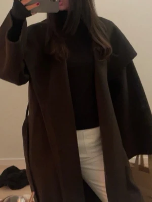 Kappa  other stories  - Säljer en stilren mörkbrun kappa med bred krage och lång passform. Jackan har en loose fit och är perfekt för dig som gillar minimalistisk och clean stil. Materialet känns tjockt och mjukt, troligen ull eller syntet. Passar dig som vill ha en enkel men snygg look.