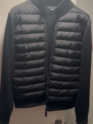 Svart pufferjacka Canada Goose - Svart pufferjacka från Canada Goose med stickade ärmar och quiltad front. Jackan har dragkedja hela vägen och en snygg logotyp på ärmen. Materialmix med polyester och stickad ull ger både värme och stil. Perfekt för dig som vill ha en clean look med premiumkänsla.äkta stl S  ett pytte litet hål vid armhålan