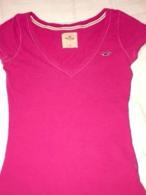 Rosa v-ringad t-shirt från Hollister - Säljer en snygg rosa t-shirt från Hollister med v-ringad hals och korta ärmar. T-shirten har en liten Hollister-logga broderad på bröstet och är gjord i mjuk bomull. Perfekt för dig som gillar enkel och stilren design.