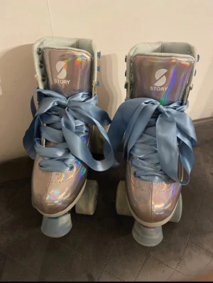 Holografiska rullskridskor från Story - Säljer ett par riktigt snygga rullskridskor från Story med holografisk yta i silver och breda ljusblå satinband som snörning. Skorna har högt skaft, rund tå och platt sula. Hjul och underrede har också en regnbågsskimrande finish som gör dem extra coola.