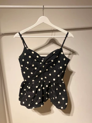 Svart prickig linnetopp Mint & Berry - Säljer en svart linnetopp från Mint & Berry i storlek 36. Toppen har vita prickar, smala justerbara axelband och en volang nedtill som ger en söt och lekfull look. Materialet är bomull och toppen är ärmlös med en lätt croppad passform.