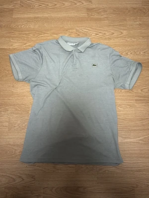 Grå pikétröja från Lacoste - Säljer en stilren grå pikétröja från Lacoste i storlek L fast passar som M. Tröjan har klassisk krage, korta ärmar och knappar framtill. Materialet är mjukt och ventilerande bomull, perfekt för en chill och snygg look. Den ikoniska Lacoste-loggan med krokodil finns på bröstet.