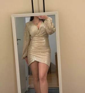 Glittrig kort klänning  - Säljer en glittrig kort klänning från Bubbleroom i en ljus champagnefärg. Klänningen har omlott-design, långärmade puffärmar och v-ringad hals. Materialet är polyester med paljetter över hela klänningen. Perfekt för att sticka ut med snyggt skimmer och trendig passform.