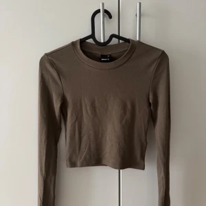 Khaki croppad långärmad topp Gina Tricot - Säljer en enkel och stilren khaki croppad långärmad topp från Gina Tricot. Toppen har rund halsringning och är tillverkad i mjuk bomull, perfekt att matcha med jeans eller kjol för en clean look.