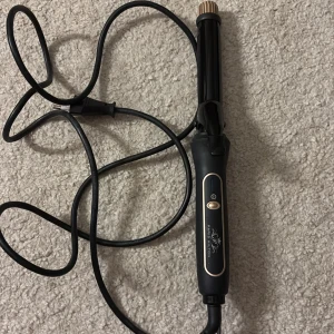 Paris Hilton Curling Iron - Elegant locktång från Paris Hilton med svart och guld design. Utrustad med enkel on/off-knapp och lång sladd för smidig användning. Produkten ser ut att vara i gott skick utan synliga defekter eller slitage. Perfekt för att skapa vackra lockar hemma.