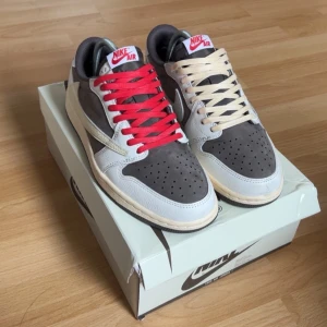 Nike Air Jordan 1 Low Travis Scott - Nike Air Jordan 1 Low Travis Scott med grå mocka, vita och beige läderdetaljer samt ikonisk omvänd Swoosh. Ena skon har röda skosnören, den andra beige. Röd broderad logga på hälen och klassisk Air Jordan-tunga. Perfekt för dig som vill sticka ut.