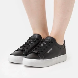 Svarta sneakers från Calvin Klein - Snygga svarta sneakers från Calvin Klein med vit platt sula och klassisk rund tå. Skorna har svarta snören, diskret logga på sidan och detaljer i metall vid snörningen. Tillverkade i skinnliknande material för en stilren look. Pris kan diskuteras.