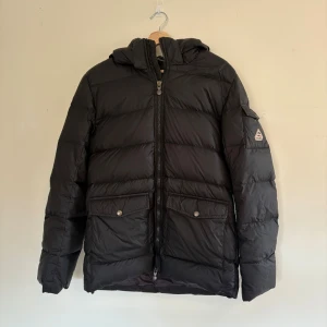Svart dunjacka från Pyrenex med huva - Svart dunjacka från Pyrenex med avtagbar  fuskpälsdetalj. Jackan har två stora fickor framtill med knappar, dragkedja hela vägen och klassisk pufferstil. Perfekt för vintern. I princip oanvänd