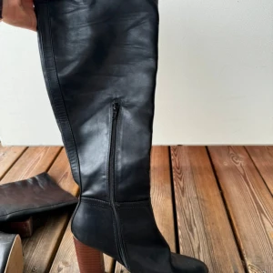 Svarta höga stövlar med klack - Säljer ett par svarta höga stövlar i skinn med snygg träklack och dragkedja på sidan. Stövlarna har en rund tå och en stilren design som passar till många olika outfits. Perfekta för dig som vill ha en klassisk look med lite edge.