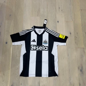 Newcastle United adidas matchtröja - Snygg Newcastle United fotbollströja från adidas med klassiska svarta och vita ränder, klubbmärke på bröstet och sponsorlogga. Tröjan har korta ärmar, gula detaljer på ärmen och är tillverkad i lätt och ventilerande material.