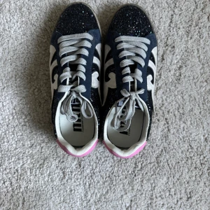 Glittriga sneakers från MTNG, strl 36 - Säljer ett par snygga sneakers från MTNG i storlek 36. Skorna är mörkblå med glitter, vita detaljer och grå snörning. Baktill har de en rosa hälkappa som ger extra färg. Komfortabel innersula och cool design med vit dekor på sidorna.