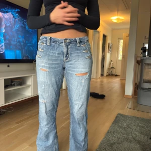 Ljusblå slitna missme jeans med knappar - Säljer ett par ljusblå jeans med flera dekorativa knappar vid fickorna och slitna detaljer framtill. Jeansen har raka ben och klassiska bakfickor med lock. Perfekta för en avslappnad och trendig look. rätt slitna, se sista bilden 