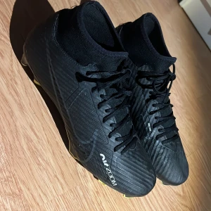Nike Air Zoom svarta fotbollsskor - Säljer ett par svarta Nike Air Zoom fotbollsskor med snörning och högt skaft. Skorna har en snygg randig textur och gula detaljer på dobbarna. Swoosh-loggan syns på sidan och materialet är syntet för lätthet och bra grepp på planen. Storlek 40,5. Ops Nike boxen får man med på köpet.