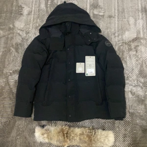 Svart dunjacka från Canada Goose L - Säljer en svart dunjacka från Canada Goose i storlek L. Jackan har huva, avtagbar pälskant och klassisk patch på ärmen. Perfekt för kalla vinterdagar med sin tjocka vaddering och stilrena look. Kommer med originalpåse och etiketter kvar.